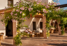 Dos personas sentadas en la terraza de un hotel rural de color blanco rodeado de árboles y flores en un día soleado, un lugar ideal para dormir cerca del Parque Natural Cabárceno.